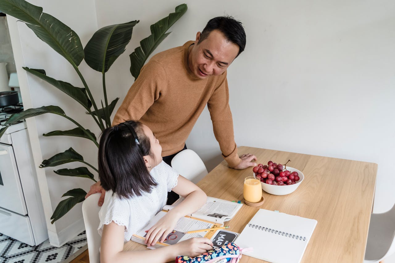 A father assists his daughter with homework at the kitchen table.