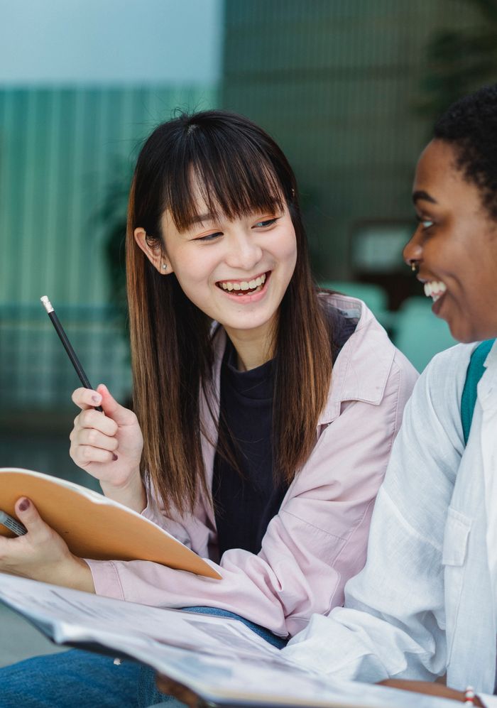 Smiling young multiracial ladies in casual outfit sitting outside with notebook and papers and speaking with each other in daytime and looking at each other
