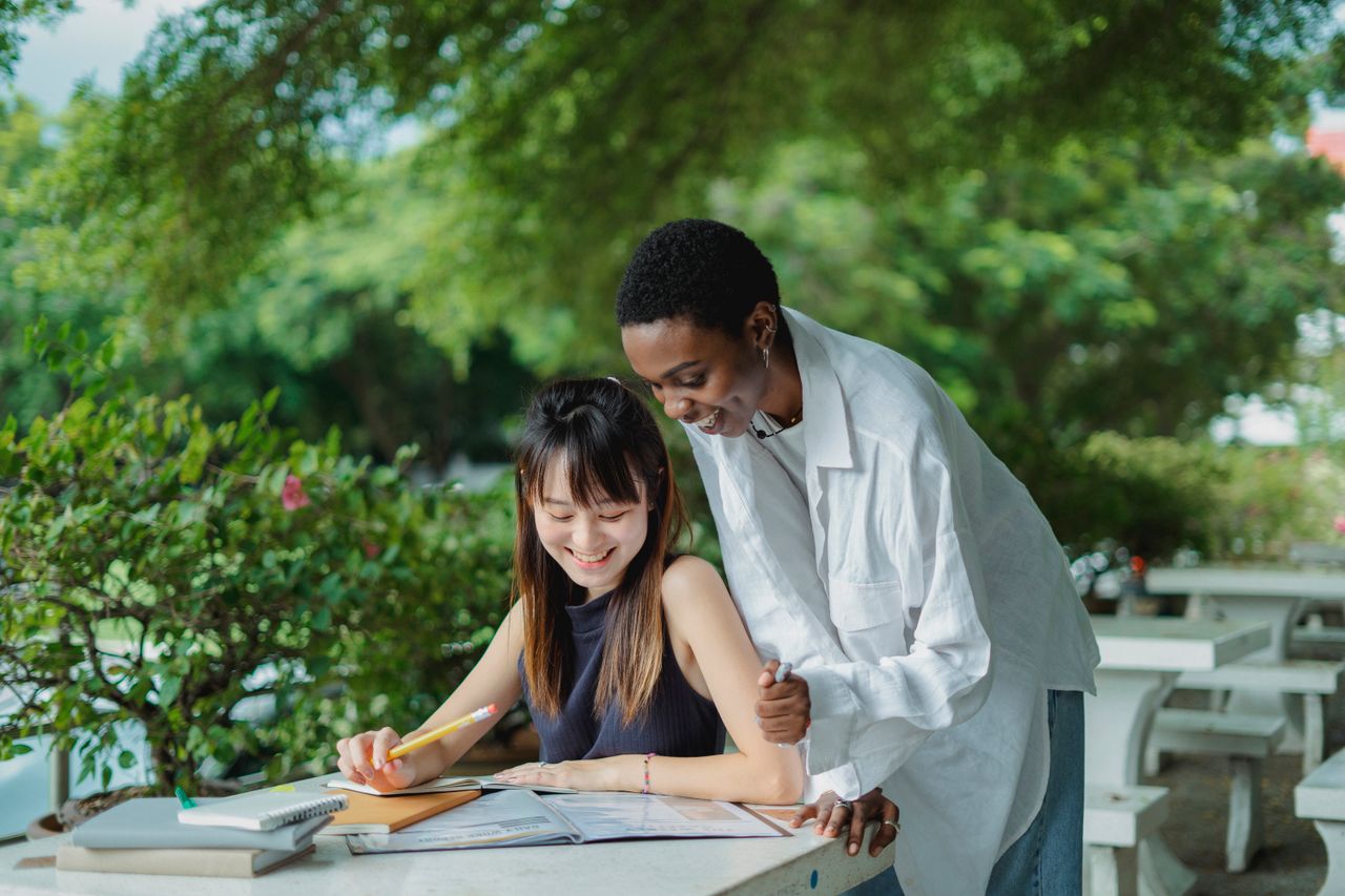 Young multiethnic women tutoring with books while spending time outdoors together and wearing casual outfit in summer day at table